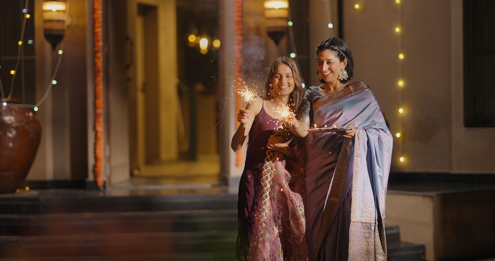 Indian Hindu happy elder family in traditional clothes play firecrackers celebrate festive season decor diya light outdoor home. Smiling mother with young adult daughter enjoy Diwali festival with older aged parent outside house