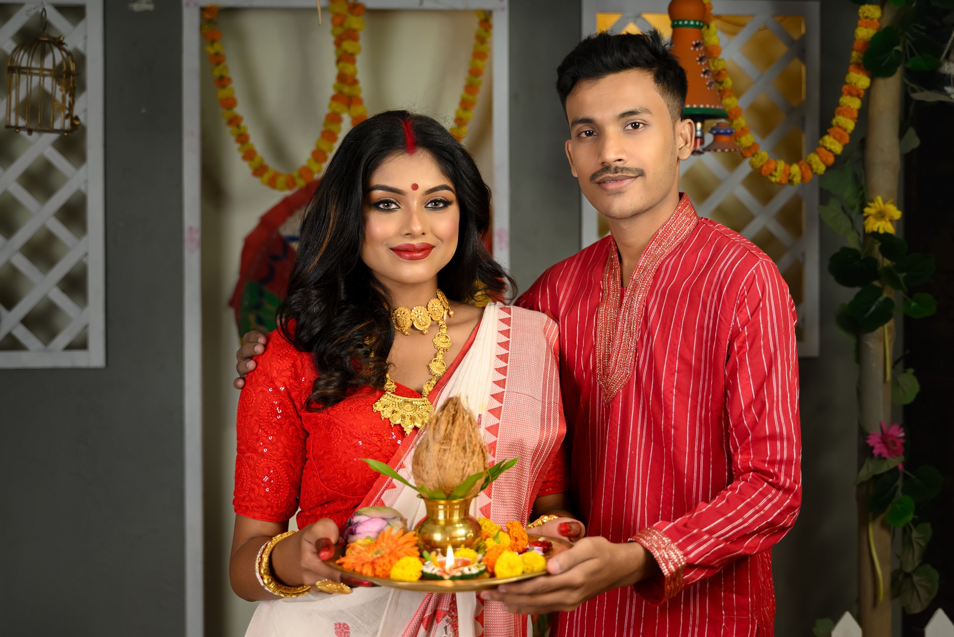 Portrait of a man dressed in a kurta pyjama stands alongside a beautiful Indian woman wearing a traditional saree, adorned with gold jewellery and bangles, holds a plate of religious offerings.